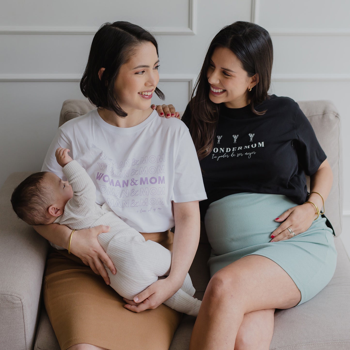 Dos mujeres sentadas. Una usando un polo color blanco que sujeta a un bebe, y otra mujer embarazada usando una falda color verde jade.