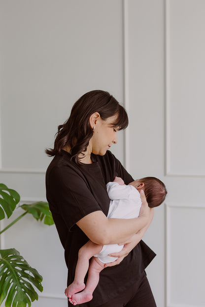 Mujer  usando un polo básico color negro. En sus brazos carga a un bebe al que le está dando de lactar.