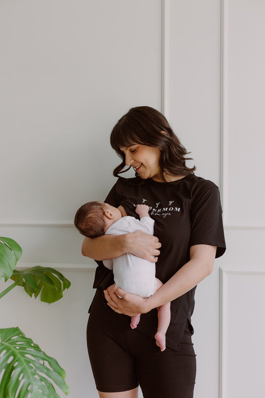 Mujer usando un polo negro. En sus brazos carga a un bebe al que le está dando de lactar.